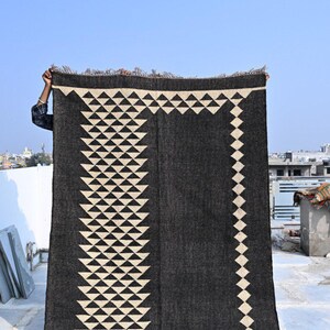 May include: A dark gray rug with a geometric design of white triangles and diamonds. The rug is rectangular with a fringed edge and is being held up against a blue sky. The design is symmetrical and covers most of the rug's surface.