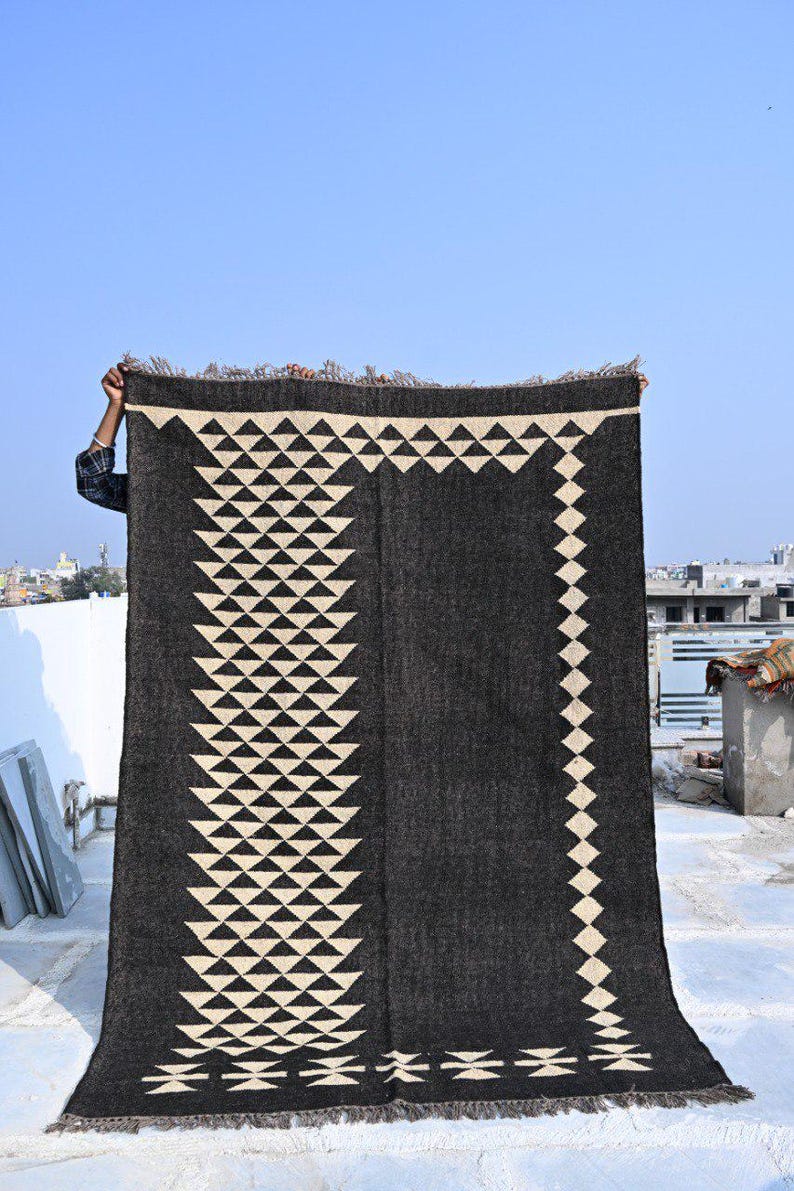 May include: A dark gray rug with a geometric design of white triangles and diamonds. The rug is rectangular with a fringed edge and is being held up against a blue sky. The design is symmetrical and covers most of the rug's surface.