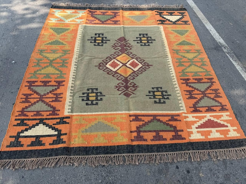 May include: A rectangular area rug featuring a central sage green panel with a geometric diamond pattern in brown, orange, and cream. The border displays repeating triangular and stepped motifs in orange, green, brown, and black, finished with fringe.