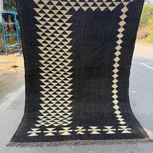May include: A large, dark gray rug with a geometric pattern of cream-colored triangles and diamonds. The rug has a fringed edge and is displayed outdoors. The design is symmetrical and covers most of the rug's surface.