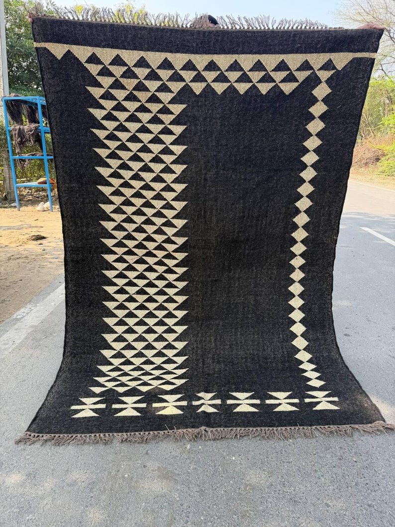 May include: A large, dark gray rug with a geometric pattern of cream-colored triangles and diamonds. The rug has a fringed edge and is displayed outdoors. The design is symmetrical and covers most of the rug's surface.