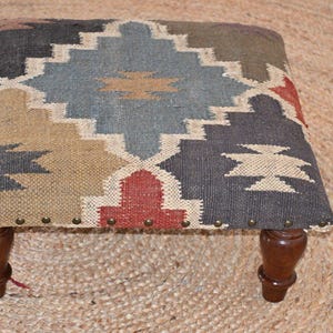 May include: A small, square footstool with a patterned top in shades of grey, beige, red, and blue. The fabric is secured with brass-coloured tacks. The footstool has four dark brown, turned wooden legs.