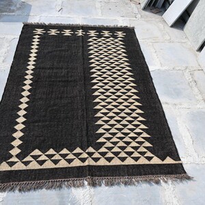 May include: A rectangular black rug with a geometric design. The rug features a border and central pattern of beige triangles and diamond shapes. The rug has a fringed edge and is displayed on a stone surface.