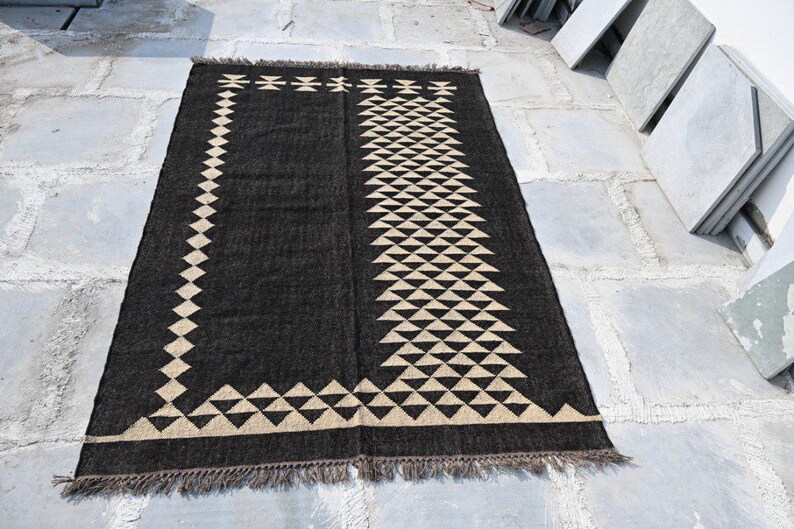 May include: A rectangular black rug with a geometric design. The rug features a border and central pattern of beige triangles and diamond shapes. The rug has a fringed edge and is displayed on a stone surface.