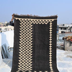 May include: A large, rectangular black rug with a geometric design. The rug features a repeating pattern of cream-colored triangles and diamonds. The edges have a fringed trim, and the rug is held up against a clear blue sky.
