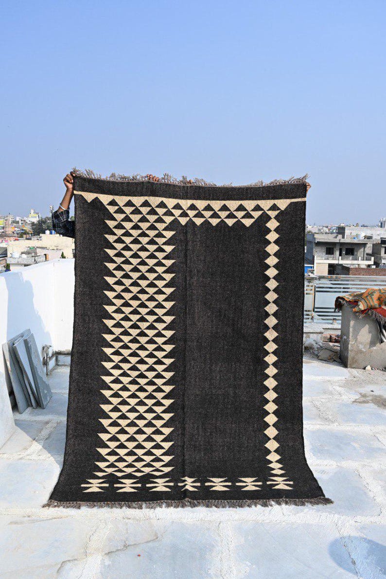 May include: A large, rectangular black rug with a geometric design. The rug features a repeating pattern of cream-colored triangles and diamonds. The edges have a fringed trim, and the rug is held up against a clear blue sky.