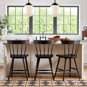May include: Three black wooden counter stools with a spindle back design sit in front of a kitchen island. The stools are in front of a window with a view of green trees.