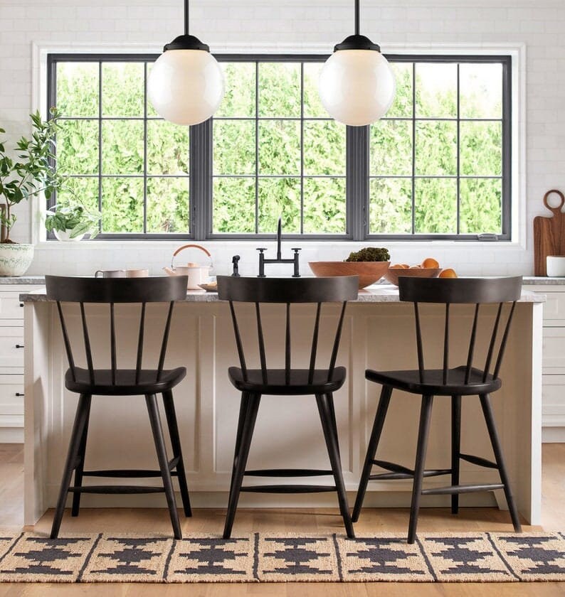 May include: Three black wooden counter stools with a spindle back design sit in front of a kitchen island. The stools are in front of a window with a view of green trees.