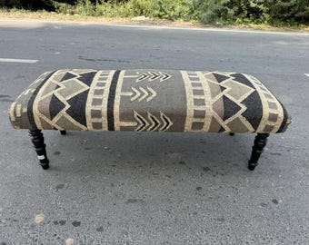 Puede incluir: Banco rectangular con una parte superior estampada y patas negras. La parte superior presenta un diseño geométrico en tonos grises, negros y beige. El banco está sostenido por cuatro patas de madera torneadas y negras, añadiendo un toque de elegancia.