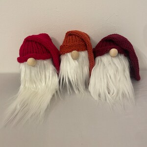May include: Three knitted gnomes with white fur beards. The gnomes are wearing red, orange and burgundy hats.