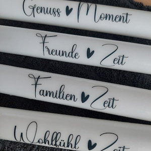 Könnte beinhalten: Vier weiße zylindrische Objekte mit schwarzem Text und Herzsymbolen. Der Text lautet "Genuss Moment", "Freunde Zeit", "Familien Zeit" und "Wohlfühl Zeit".