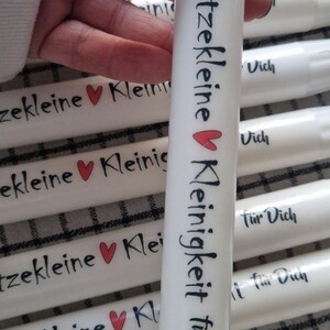 Könnte beinhalten: Weiße Kerzen mit schwarzer Schrift und roten Herzakzenten. Der Text auf den Kerzen lautet "klitzekleine Kleinigkeit für Dich" und "klein". Die Kerzen sind zylindrisch und haben einen Docht oben.