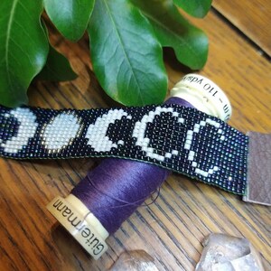Puede incluir: Un brazalete de cuentas con un dise&ntilde;o de lunas en blanco y negro. El brazalete est&aacute; sobre un carrete de hilo morado y una superficie de madera. Hay dos cristales transparentes al lado del carrete.