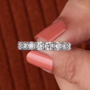 May include: A silver eternity ring with multiple round-cut, clear gemstones. The ring is held against a warm, orange-brown background. The gemstones are set closely together, creating a continuous sparkle.