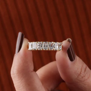 May include: A gold band ring featuring a row of rectangular-cut, clear gemstones. The ring is held between two fingers against a blurred, reddish-brown background. The gemstones are set closely together, creating a continuous sparkle.