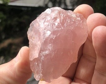 May include: A raw, translucent rose quartz crystal with a pale pink hue and an irregular, jagged shape. The stone features a crystalline texture with visible facets and natural inclusions, displaying a soft, light-reflecting surface throughout.