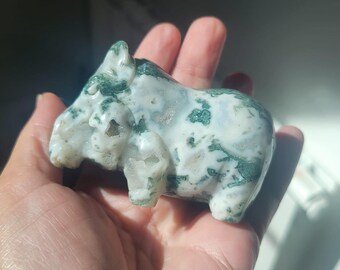 Puede incluir: Pequeña figura de piedra tallada de un hipopótamo. La escultura es blanca con inclusiones verdes y grises, lo que le da un aspecto natural. El hipopótamo está de pie, con las patas y el cuerpo claramente definidos. La figura se sostiene en una mano.