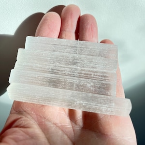 Può includere: Una pila di cristalli di selenite traslucidi e rettangolari. I cristalli sono impilati in una formazione a strati, con striature visibili. La selenite ha un aspetto bianco e morbido. È tenuta in una mano.