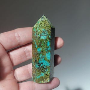 Può includere: Un obelisco di cristallo turchese e verde con la parte superiore a punta. La pietra presenta un motivo screziato di tonalità blu, verdi e marroni. Il cristallo è tenuto in mano.
