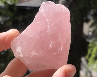 May include: A raw, unpolished chunk of rose quartz crystal with a translucent, pale pink hue. The stone features an irregular, jagged shape with natural facets and internal fractures that catch the light, displaying a soft, cloudy appearance.