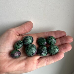 Peut inclure: Huit pierres polies de rubis dans du zoïsite sont présentées dans une main. Les pierres ovales ont une base vert foncé parsemée de noir et de taches de rubis rouge-rosé. Le fond est un mur blanc uni.