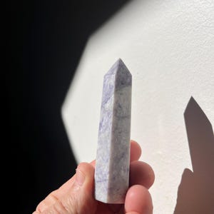 Peut inclure: Une pointe de cristal violet clair avec un dessus facetté et pointu. Le cristal est tenu dans une main sur un fond blanc avec une ombre sombre. Le cristal a un aspect marbré avec des stries blanches et violettes.