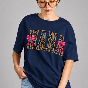 Könnte beinhalten: Ein marineblaues T-Shirt mit dem Wort "NANA" in einem Leopardenmuster-Design, umrandet von Gold. Auf beiden Seiten des Wortes befinden sich rosa Schleifen. Das Model trägt eine Jeans und eine Brille.