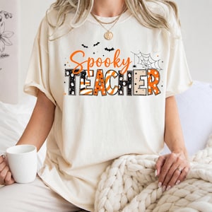 Könnte beinhalten: Ein cremefarbenes T-Shirt mit dem orangefarbenen Schriftzug Spooky und dem Wort TEACHER in schwarzen, orangefarbenen und weißen Blockbuchstaben. Das Design zeigt schwarze Fledermäuse, ein Spinnennetz, kleine Sterne und Geistersilhouetten.