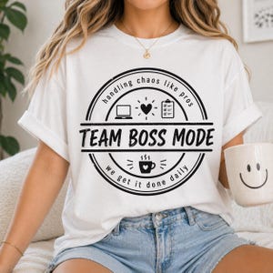 May include: White t-shirt with a black circular graphic that reads "handling chaos like pros" and "Team Boss Mode." The graphic includes a laptop, heart, checklist, and coffee cup. A white mug with a smiley face is held in the hand.
