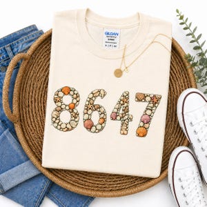 Könnte beinhalten: Cremefarbenes Sweatshirt mit den Zahlen "8647", die aus Muscheln und Seesternen geformt sind. Das Shirt liegt auf einem geflochtenen Korb mit blauen Jeans und weißen Turnschuhen. Eine goldene Halskette mit Anhänger ist ebenfalls zu sehen.
