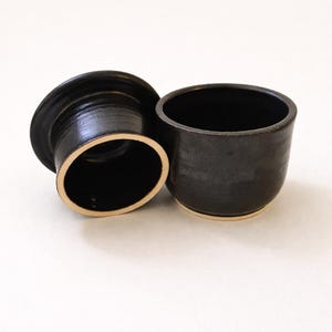 Peut inclure: Un beurrier en céramique noire avec un couvercle et un bol séparé. Le plat a une finition mate avec un bord de couleur claire. Le couvercle est de forme ovale et repose à côté du bol, qui a une forme arrondie.