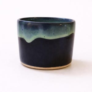 Peut inclure: Une tasse en céramique faite à la main, avec une base bleu foncé et un dessus vert clair. La tasse a une finition lisse et brillante et une forme cylindrique simple. Le fond de la tasse est de couleur naturelle non émaillée.