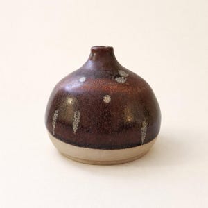 Könnte beinhalten: Kleine, handgefertigte Keramikvase mit rundem Körper und schmalem Hals. Die Vase hat eine satte, dunkelbraune Glasur mit helleren, gesprenkelten Akzenten und einem beigefarbenen Sockel. Ein dekoratives Stück.