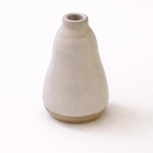Peut inclure: Un petit vase en céramique en forme de poire, avec un corps beige clair et une base contrastante de couleur beige clair. Le vase a une ouverture étroite en haut et une finition lisse et mate. Le design est simple et minimaliste, adapté pour les petites fleurs.