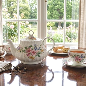 Puede incluir: Un juego de té con motivos florales, que incluye una tetera, una taza y un platillo, junto con una azucarera y una lechera plateadas. Hay scones y macarons en un plato. La escena está ambientada en una mesa de madera cerca de una ventana con vistas al jardín.