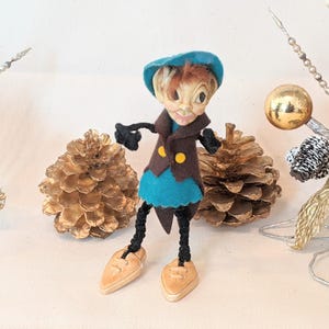 Puede incluir: Un muñeco de elfo de Navidad de fieltro vintage con un sombrero azul, una chaqueta marrón y una falda turquesa. El elfo tiene una cara pintada con cabello castaño y ojos negros. El muñeco está de pie sobre una superficie blanca con piñas doradas y una decoración de árbol de Navidad plateada y dorada.