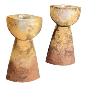 以下が含まれることがあります： 2つの抽象的なキャンドルホルダー。それぞれ、小さな穴のある丸みを帯びた上部と円錐形のベースがあります。色のグラデーションは金色から茶色に変化します。キャンドルホルダーはテクスチャード加工された素材で作られています。