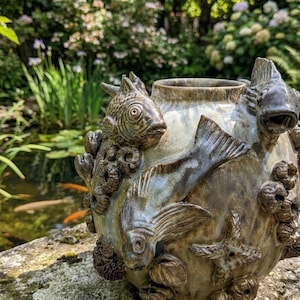 Può includere: Un vaso in ceramica rotondo con smalto maculato marrone e grigio, caratterizzato da pesci scolpiti in tre dimensioni, stelle marine e texture circolari simili a cirripedi applicate sulla superficie esterna del contenitore.