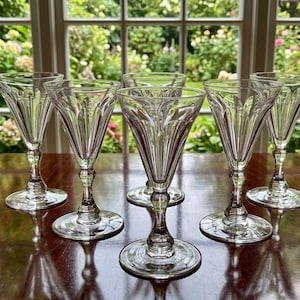 Op de afbeelding: Een set van vijf heldere glazen op voet met een uitlopende kelk voorzien van verticale geëtste lijnen en een decoratief patroon bij de rand. Elk glas heeft een slanke steel en een ronde voet, gerangschikt op een houten oppervlak.
