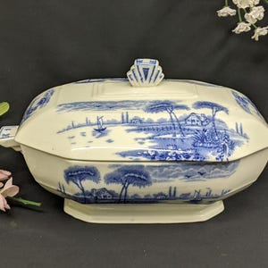 May include: A white and blue porcelain serving dish with a lid. The dish features a blue and white floral design with a cottage and trees. The lid has a decorative finial.