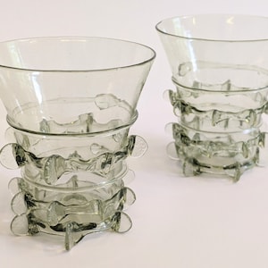 Puede incluir: Dos vasos para beber de vidrio transparente con un diseño único. Los vasos tienen una parte superior ancha y acampanada y una base estrecha. La base de cada vaso está decorada con una serie de anillos de vidrio que se han moldeado en un patrón decorativo.