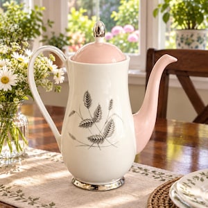 Puede incluir: Una cafetera de cerámica con cuerpo color crema y tapa y pico de color rosa suave. El frente está decorado con una ilustración botánica negra de espigas de trigo. Incluye un asa, una base y un remate superior en tono plateado.