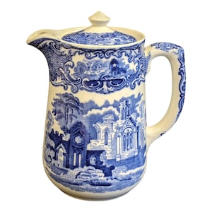 May include: A blue and white ceramic jug with a lid. The jug has a detailed blue landscape design featuring buildings and trees. The handle and spout are white. The jug is likely vintage or antique.