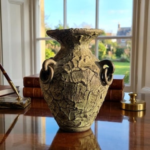 Peut inclure: Un vase en céramique avec une surface texturée et craquelée dans des tons de vert et de brun sourds. Le récipient présente un bord large et évasé ainsi que deux anses circulaires proéminentes fixées sur les côtés, lui donnant un aspect vieilli.