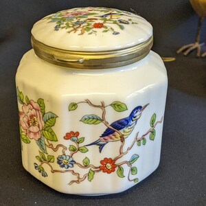 Puede incluir: Un tarro de porcelana blanca con tapa dorada y diseño floral. El tarro presenta un pájaro azul y amarillo posado en una rama con flores rosas y rojas.