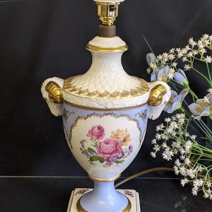 May include: A vintage porcelain table lamp with a floral design. The lamp features a white and gold top, a light blue base, and a square base with floral accents. The lamp's body is decorated with pink roses and other colorful flowers.