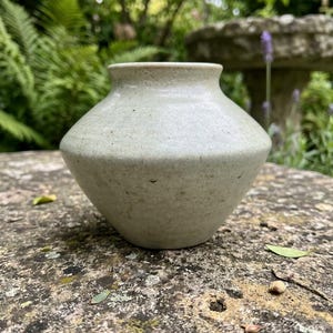 May include: A small, wide-bodied ceramic vase with a speckled, off-white matte glaze. The vessel features a tapered neck, a circular opening, and a distinct angular shoulder that transitions into a narrow, rounded base with a smooth finish.