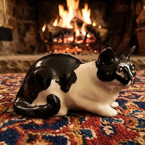 May include: A ceramic cat figurine in a sitting position, with a black and white color scheme. The cat has yellow eyes and is placed on a patterned rug. A fireplace with a burning fire is in the background.