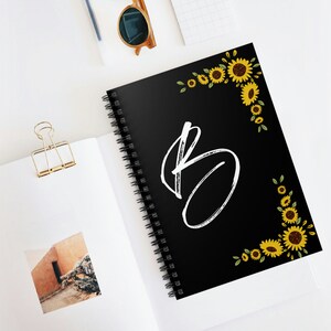 May include: Black spiral notebook with a white letter "B" and a sunflower border. The notebook is lying on a white surface.