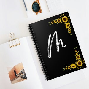 Puede incluir: Cuaderno espiral negro con una letra "M" blanca y un borde de girasoles amarillos sobre fondo negro.